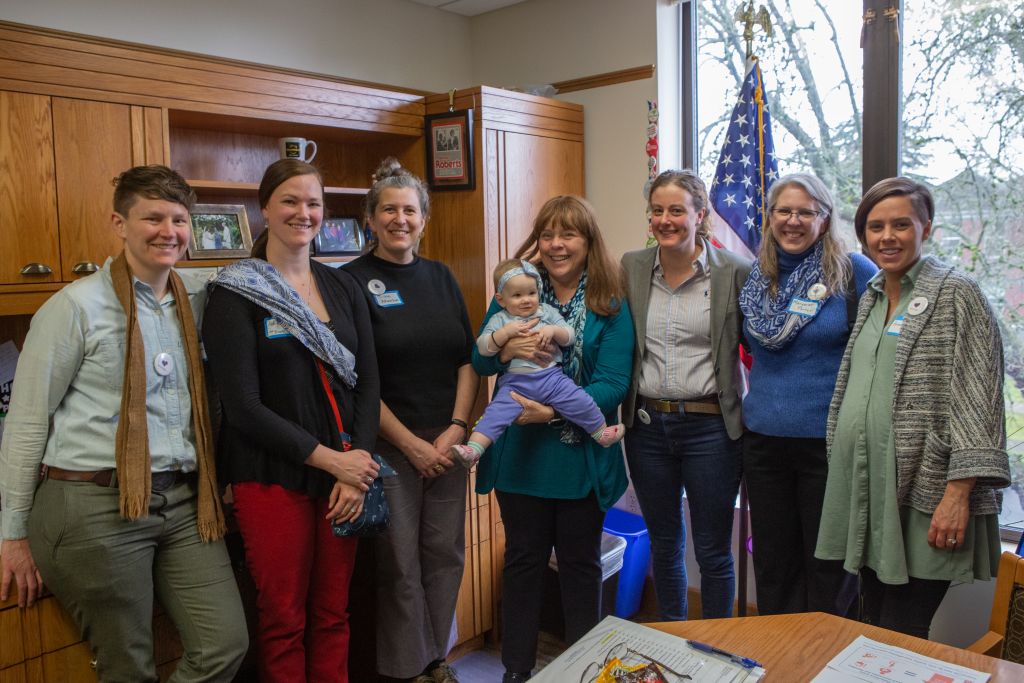 Midwives and mothers stand in a row with Representative Sheri Schouten, who holds a baby. They are in Representative Schouten's office at the Oregon Capital. 